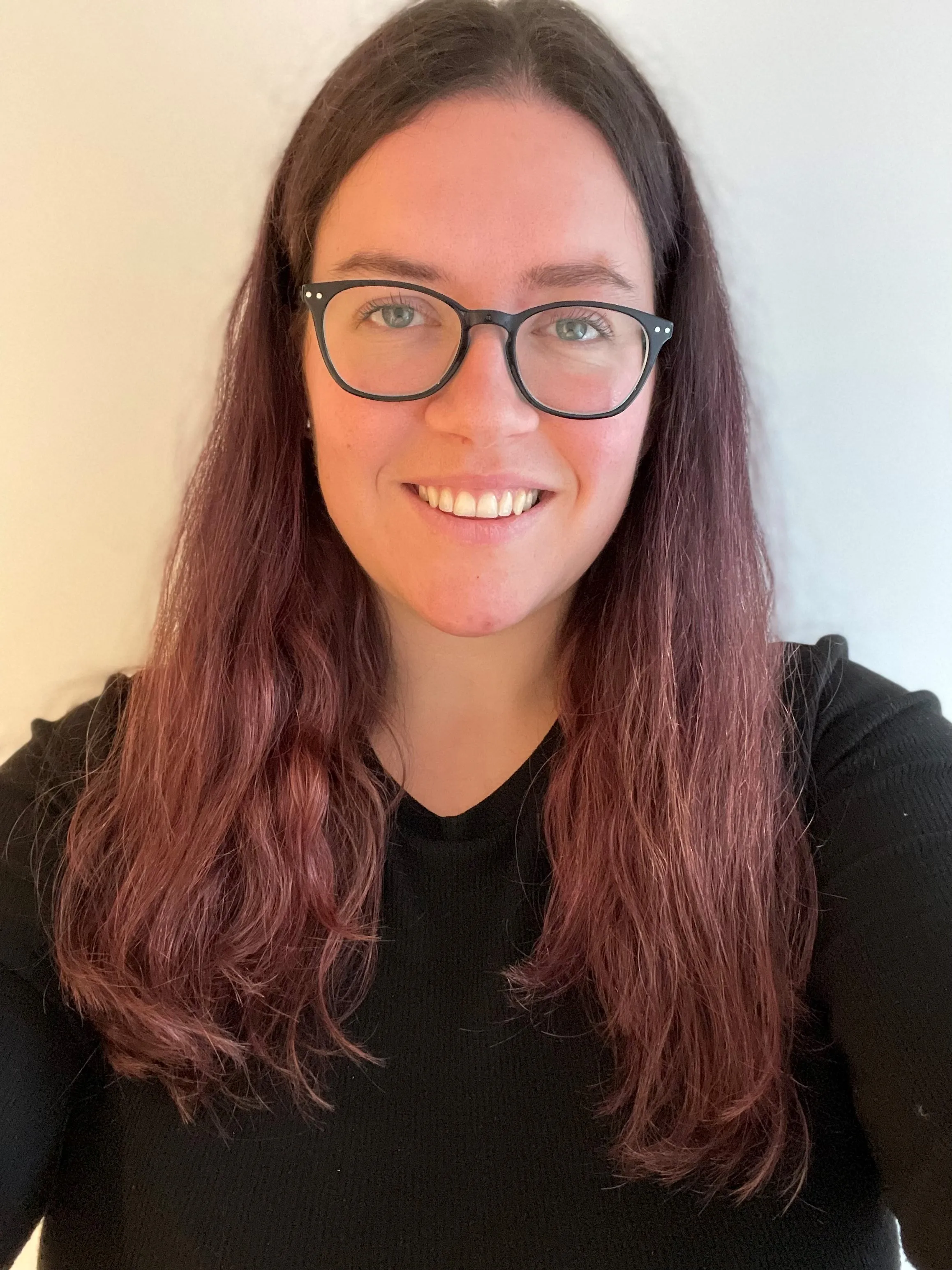 Alison is smiling and looking directly at the camera, her hair is flowing over her shoulders. She has black rounded glasses and is wearing a black top.